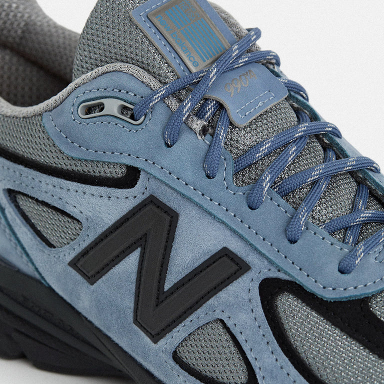 The revival of the New Balance 990 v4 after 8 years, featuring gray mesh with blue suede overlays and black details. Made in the USA with EnCap cushioning for comfort.