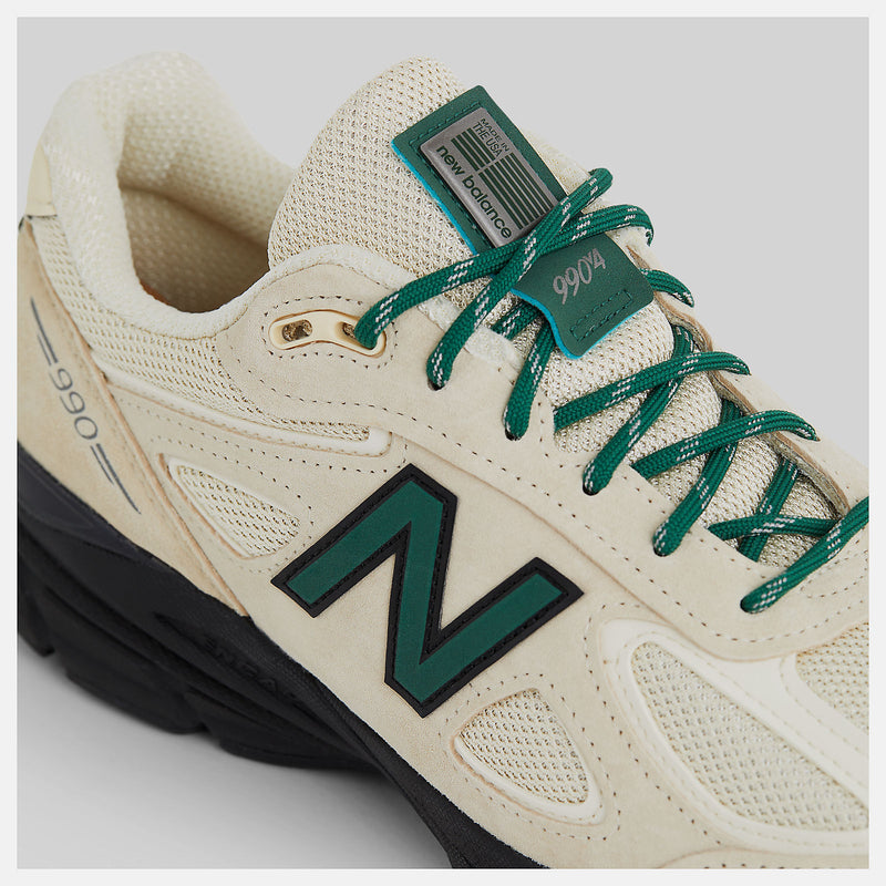 This iconic running shoe showcases the triumphant return of the New Balance 990 v4 after an 8-year hiatus. It features a light beige mesh upper adorned with suede overlays, complemented by black and dark green accents. 